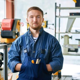 Waist up portrait of young mechanic posing looking at camera standing in industrial workshop, copy space